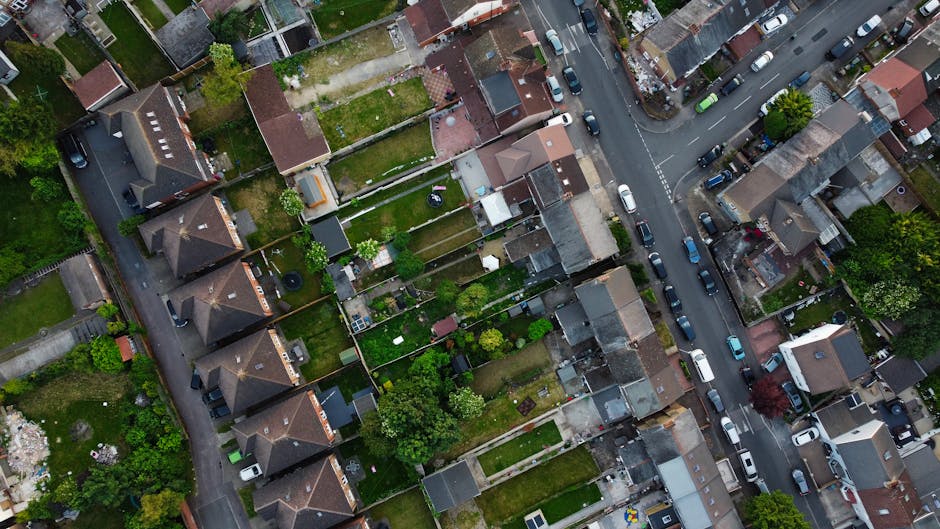 A quiet residential street on a sunny day featuring a row of traditional terraced houses with brick facades and pitched roofs. Each property has well-maintained front gardens filled with lush, green h